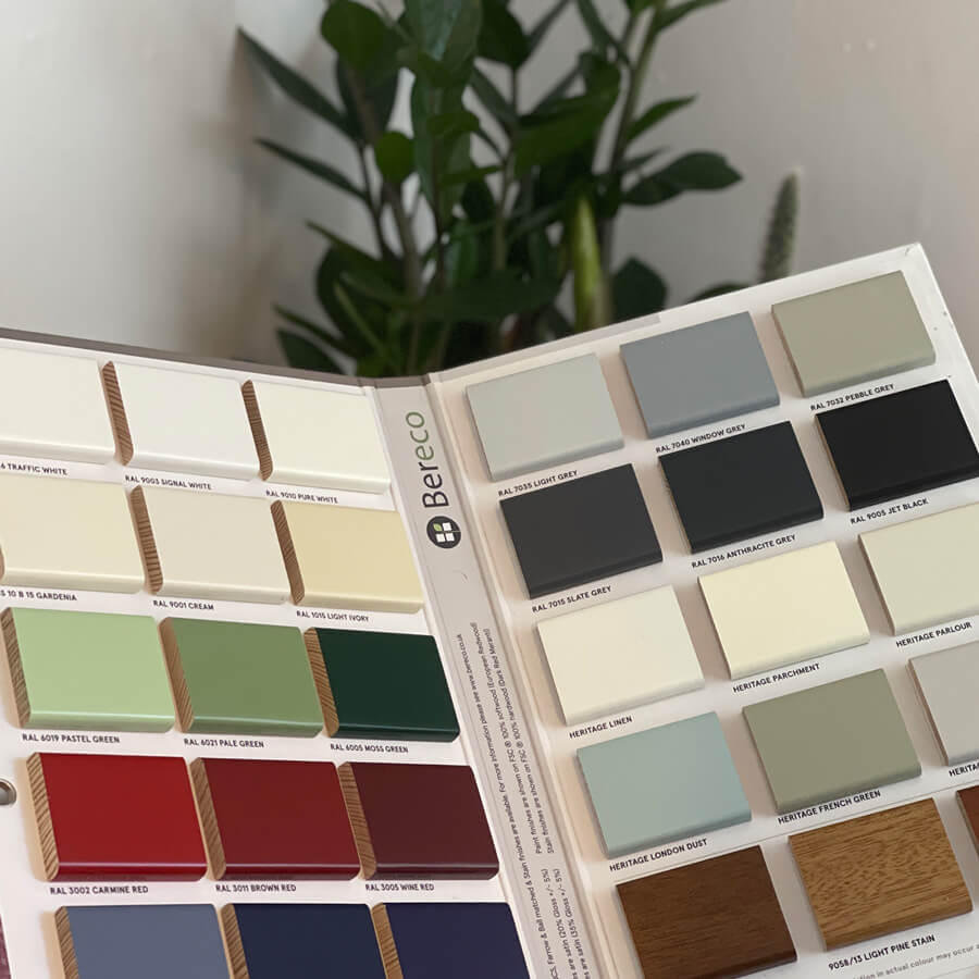 A color sample book open to various paint swatches, labeled with names and codes, sits on a wooden surface. A green plant is in the background. Text: "Bereco".