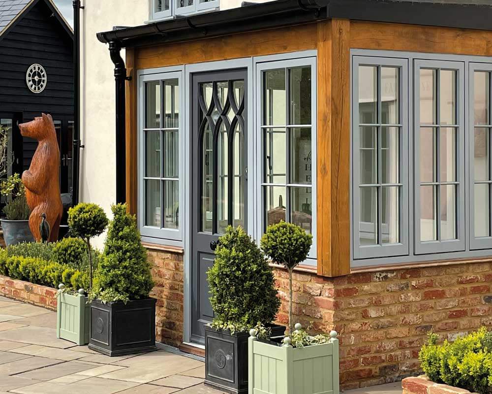 A wooden bear statue stands beside a timber windowed porch with light blue frames, surrounded by boxwood shrubs in planter boxes, situated in front of a brick base and beige house.