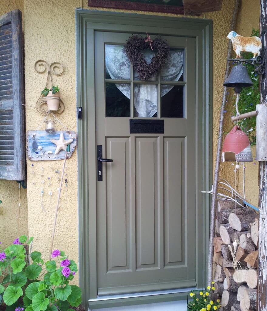 Green timber door adorned with a heart-shaped wreath stands closed against a yellow house, flanked by potted plants, hanging decor, and a stack of firewood.