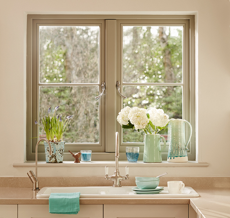 A kitchen sink sits below a window with potted flowers on the sill. Surrounding utensils include a turquoise jug, blue and white cups, a bowl, and a green towel draped over the sink. Forest view through the window.