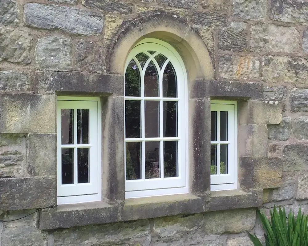 A white-trimmed, medieval-style timber arched window with two smaller rectangular windows is set in a rustic, weathered stone wall. Green foliage is partially visible at the window's base.