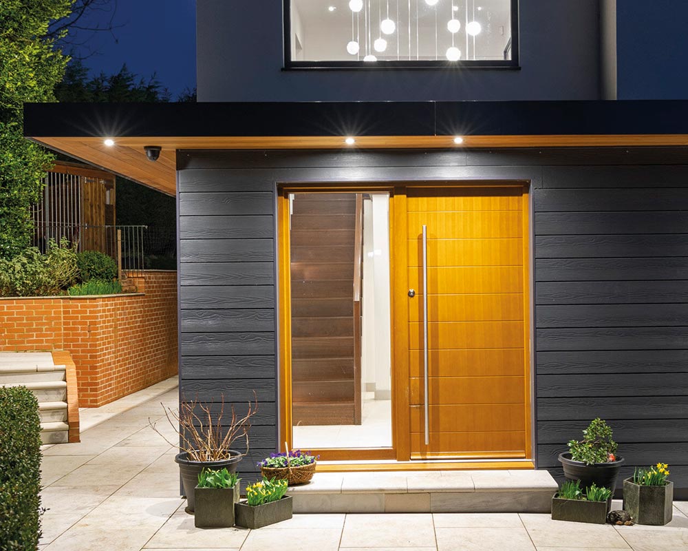 An illuminated wooden front door with a tall handle leads into a modern home; the exterior features dark siding, potted plants, and a brick wall in a nighttime setting.
