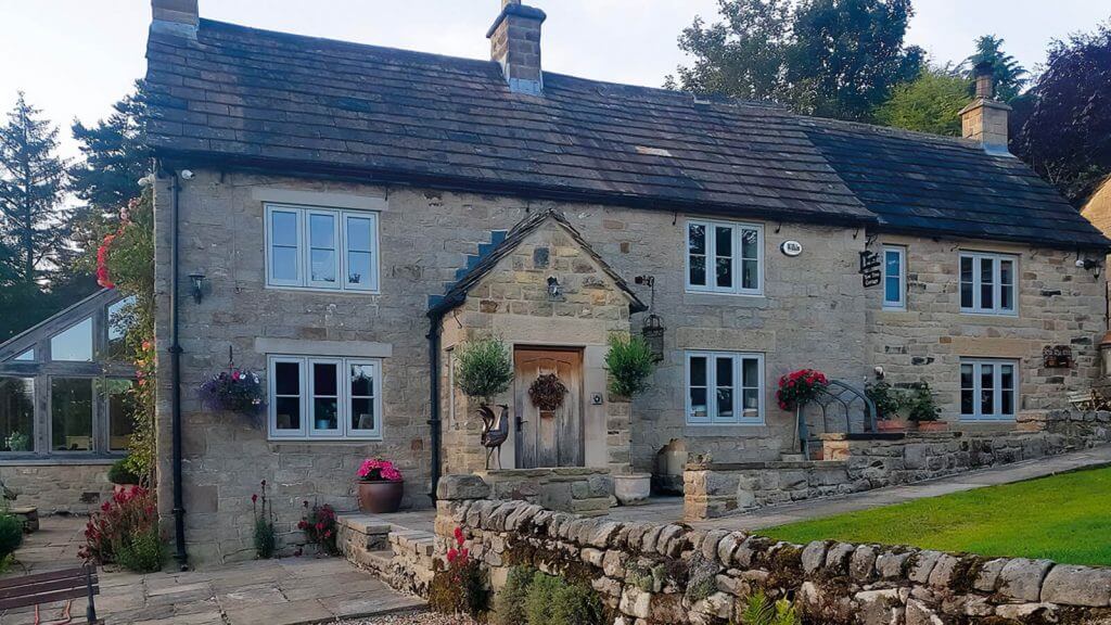 Stone cottage with a slate roof and white-framed windows, adorned with potted plants and hanging flowers. It is surrounded by a stone wall and a well-maintained garden, with trees in the background.
