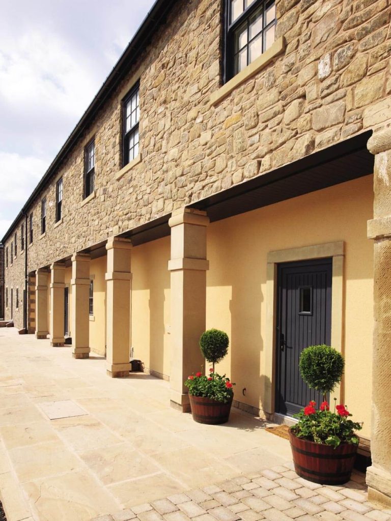 Stone building with a row of windows and doors; potted plants with topiary and flowers sit on a tiled pathway under a sunny, partly cloudy sky.
