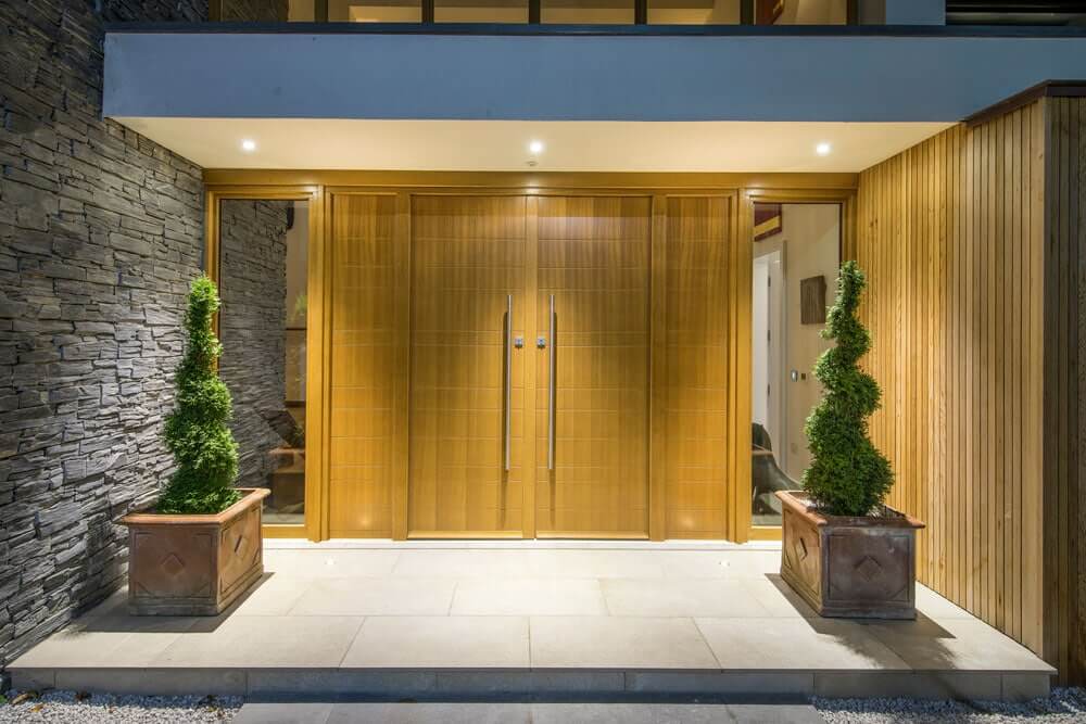 A double wooden door stands closed, flanked by two spiral-shaped topiary plants in square planters, with a stone wall on the left and a wooden wall on the right, all illuminated by recessed lights.