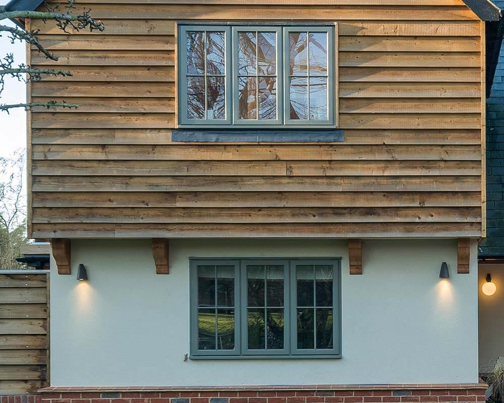 A two-story building facade features wood siding with a window on the upper level and two wall lights flanking a lower level window. Light trees are visible in the background.