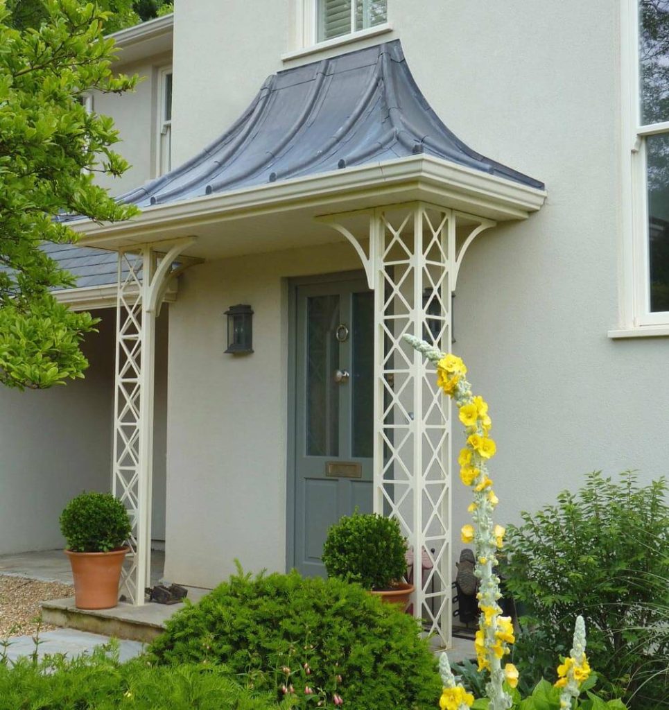 A grey door with a traditional lamp and a curved awning supported by lattice columns, flanked by potted plants, surrounded by lush greenery and yellow flowers in front of a pale-colored house.