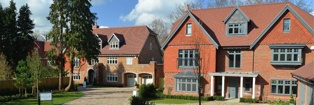 Two large, red-brick houses with white-framed timber windows and brown roofs stand side by side on a sunny day, surrounded by green lawns, trees, and a stone-paved driveway.