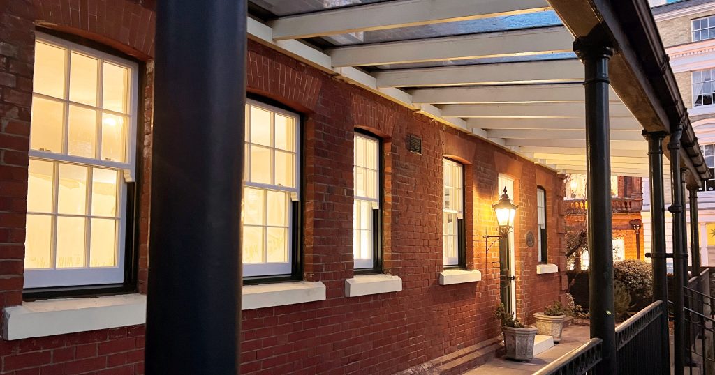 A row of warmly lit windows and a lamppost glow along a red-brick building's covered walkway, with railings and nearby greenery enhancing the nighttime urban scene.